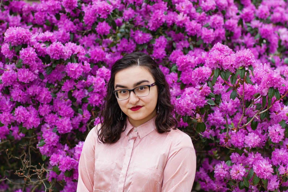 Mona Jahani, SLU theatres newest costume designer, appears in front of a bursting tree of pinkish-lavender flowers. 