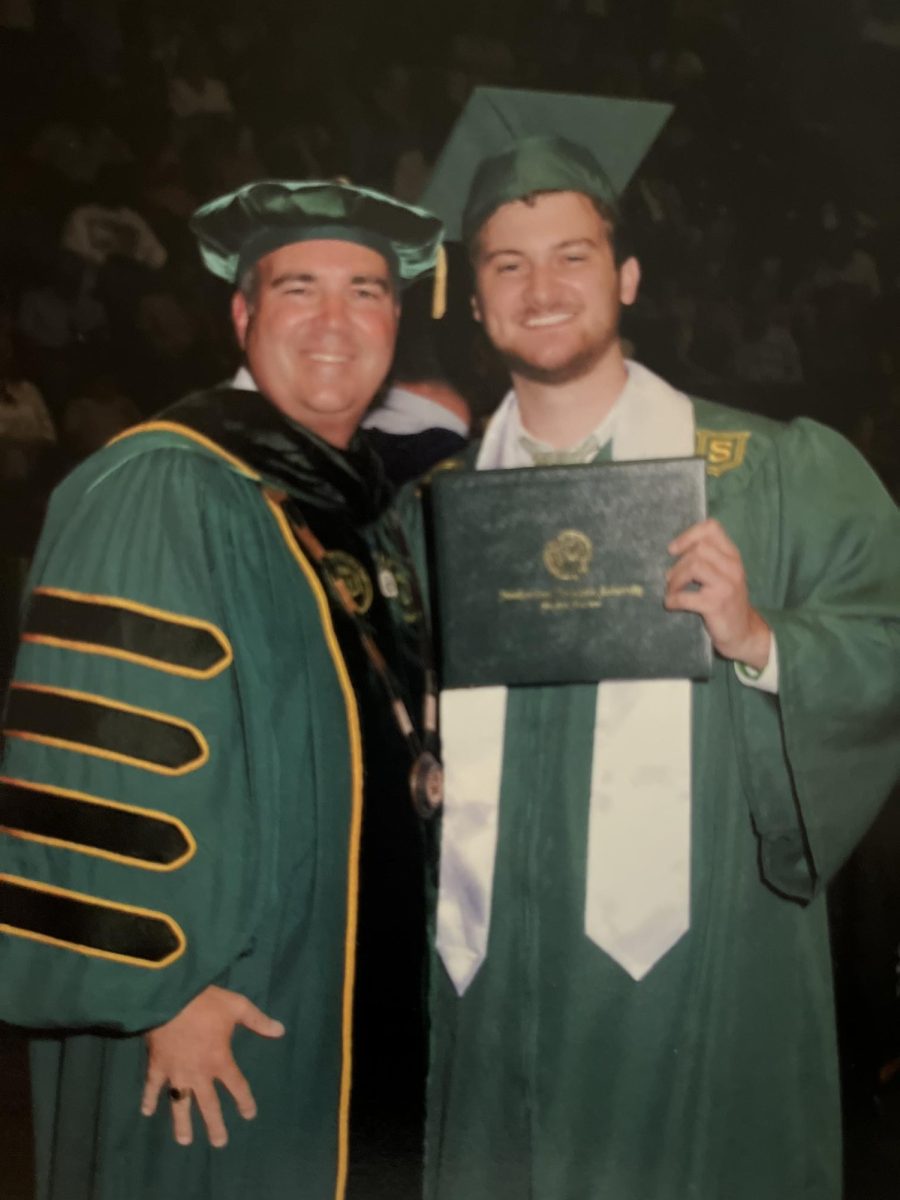 A younger man in a green graduation cap and gown smiles, holding a diploma, next to an older man in a green academic robe.