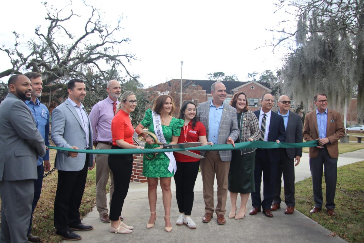 People stand in front of a tree and pose in front of a ribbon with large scissors, signaling a ribbon cutting.
