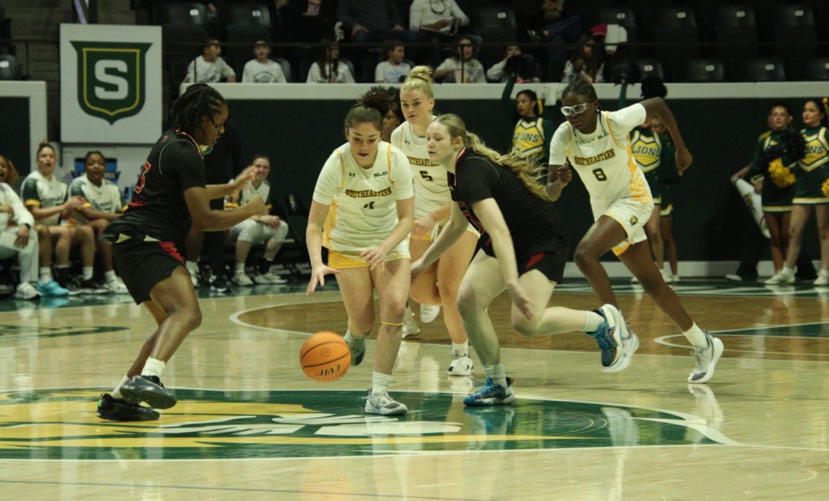 A girl dressed in white dives for the ball in the middle of the basketball court to keep it away from the girls dressed in black and red.