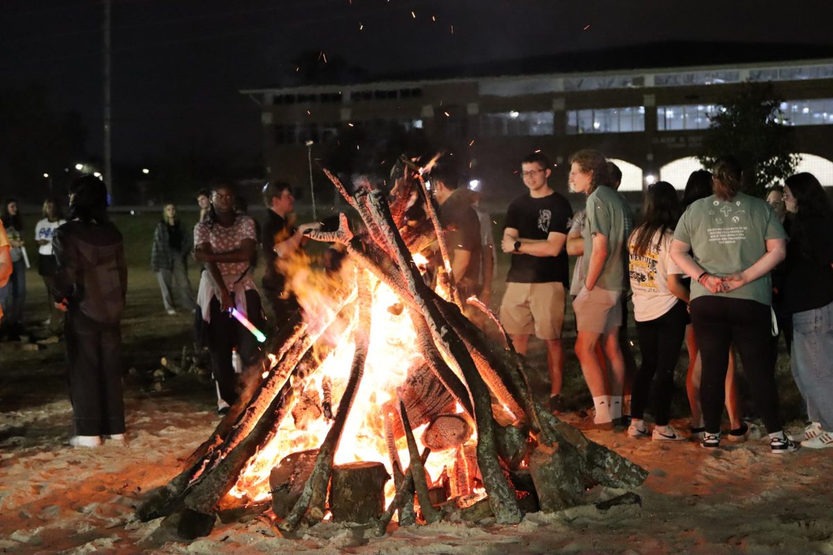 Young people stand around a fire being kindled by a bunch of wood and large sticks.