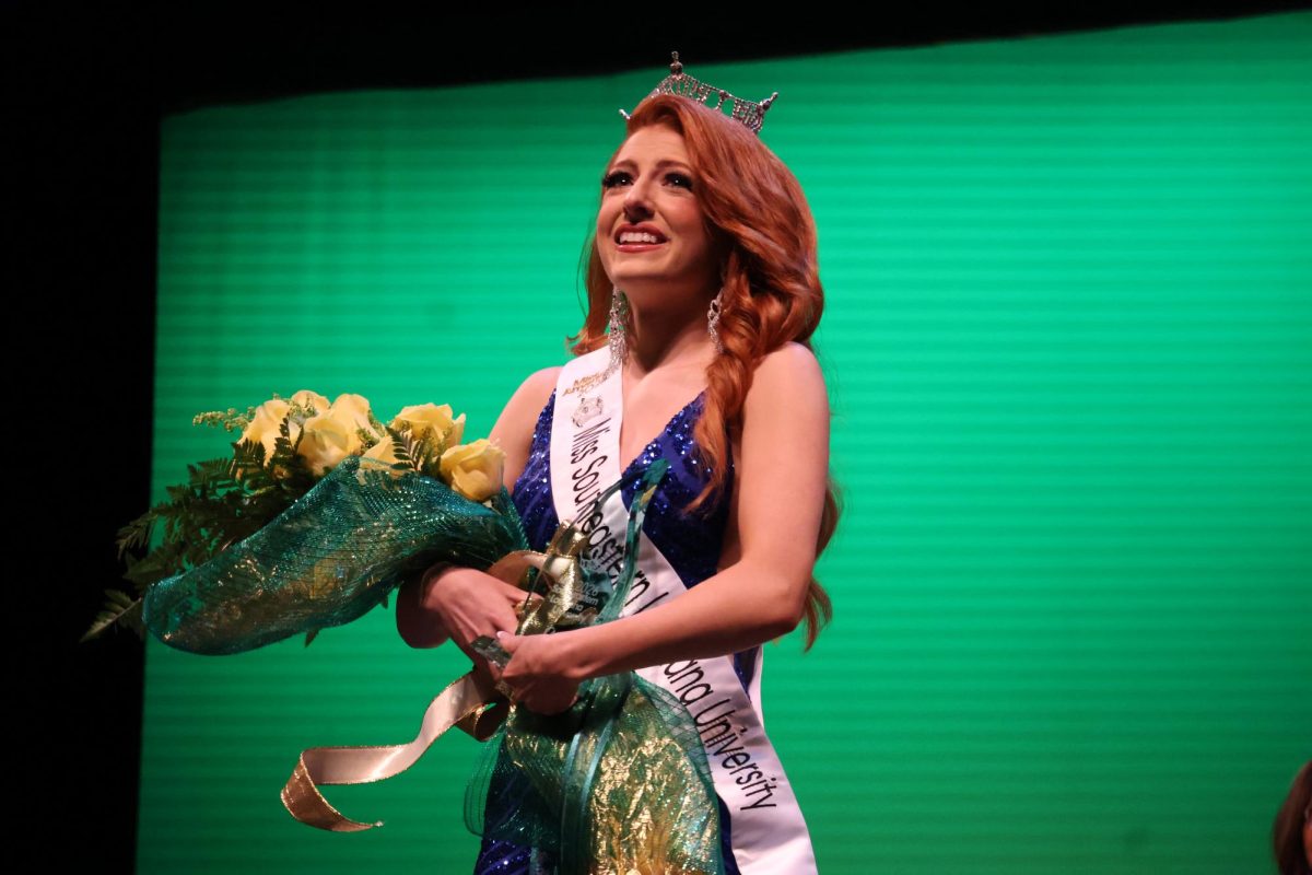 A woman in a blue dress, a crown and a sash holds a bouquet of bright yellow flowers.