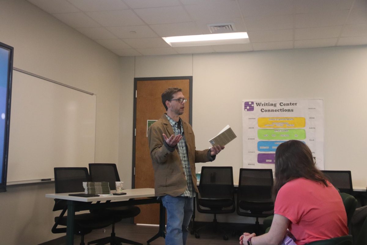 A man holds a book and talks in a room. A person in a pink shirt is in frame, listening.