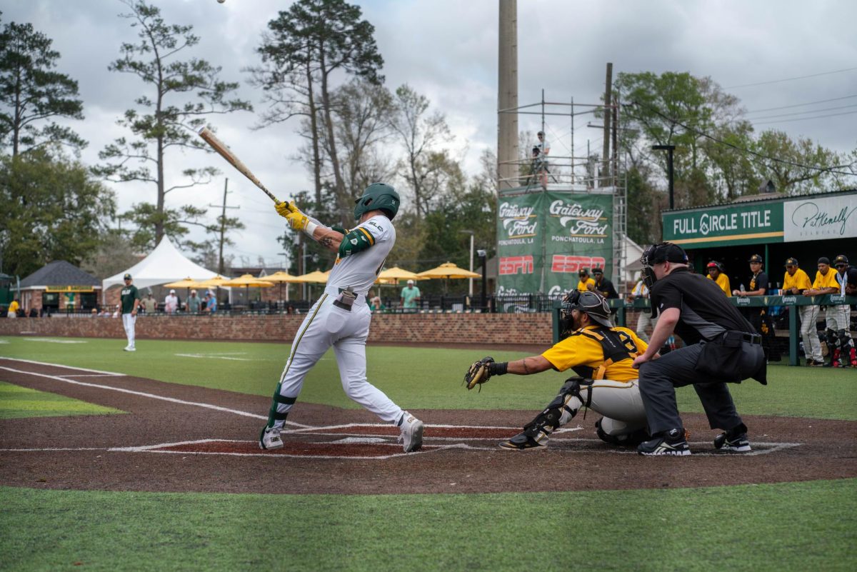 Baseball player in white swings his bat in front of a catcher with a yellow uniform and the umpire dressed in black.