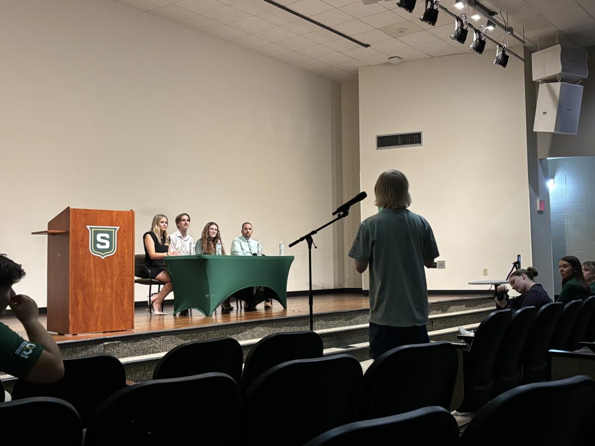 A stage with people sitting and listening to a person speaking into a mic.